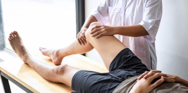A man wearing a grey shirt and black shorts lying on a massage table while a therapist in a white coat inspects their knee