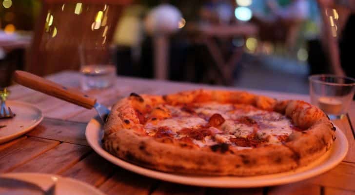 A woodfired pizza being placed on a plate with a pizza peel, on a wooden table with glasses in a restaurant