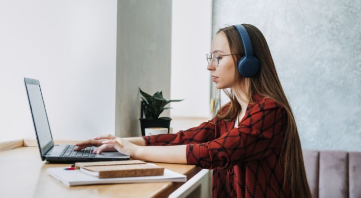 A woman wearing headphones sits at a desk and uses a laptop