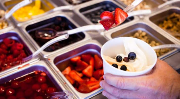 A hand holding a cup of frozen yogurt over a selection of toppings