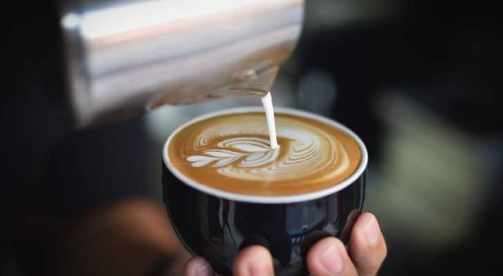 A barista holds a cup of fresh coffee and creates a pattern by pouring milk into it