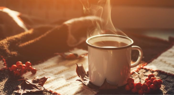 A steaming cup of coffee on a blanket surrounded by autumn leaves and spices