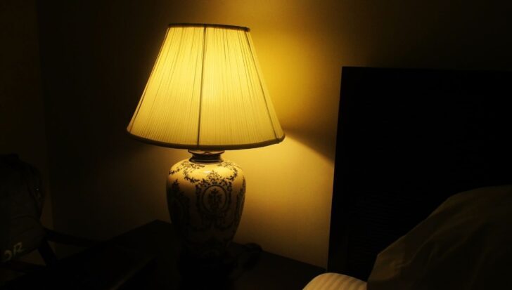 A bedside lamp with a decorated base and a lighted lampshade in a dimly lit room