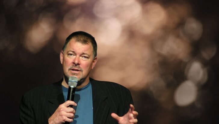 Jonathan Frakes holding a microphone, speaking with a blurred background