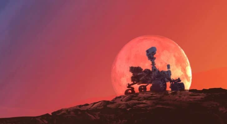 A Mars Rover sitting atop a hill on Mars with a red sky behind it