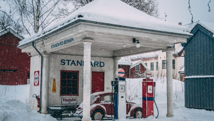 Snow-covered vintage gas station with an old red car parked, surrounded by snow and trees