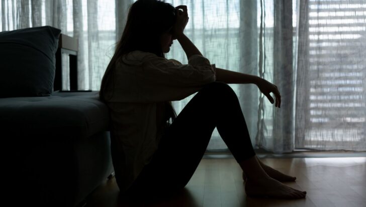 Silhouette of a woman sitting on the floor with her head resting on her hand, next to a couch in a dimly lit room