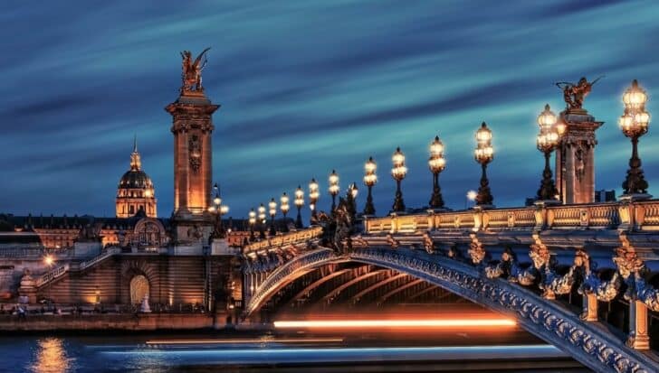 Illuminated Alexander III Bridge in Paris with ornate lamp posts and a lighted dome in the background