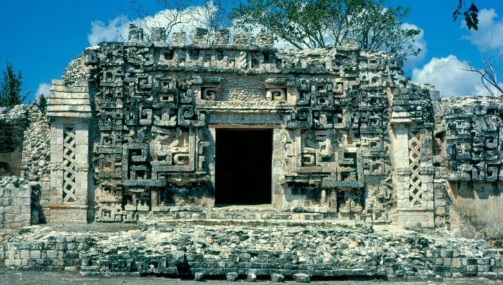 Ancient Maya temple with intricate stone carvings and a large central doorway, set against a backdrop of trees and blue sky