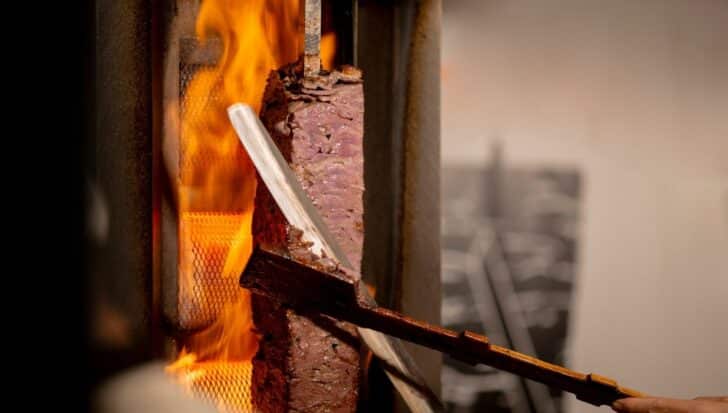 Meat being cooked on a vertical rotisserie near an open flame