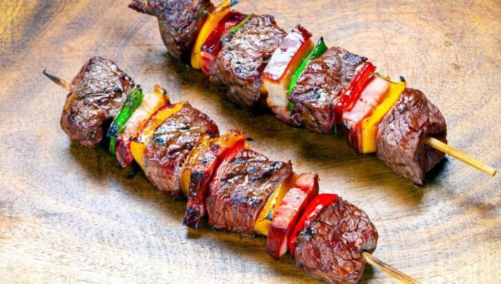 Two skewers with grilled meat and colorful vegetables on a wooden surface