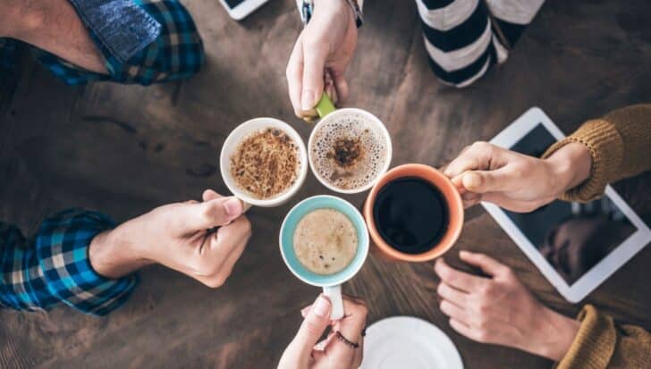 Four people holding cups of coffee over a table with a tablet on it