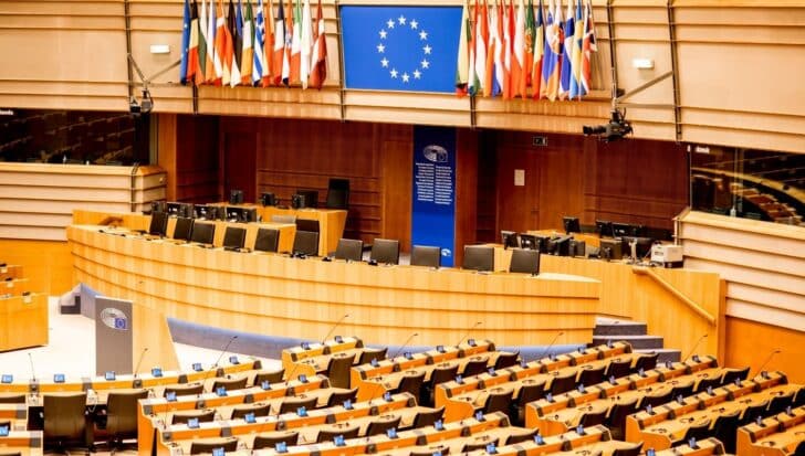 Interior of the European Parliament chamber with rows of seats, desks, and a large display of the EU flag surrounded by member state flags