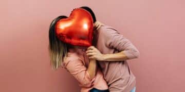 Two people holding a red heart-shaped balloon in front of their faces in a playful gesture, both wearing pink tops against a pink background