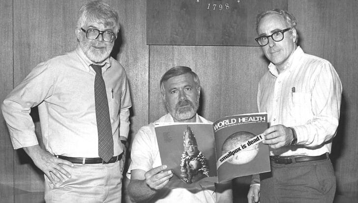 Three men stand together holding a World Health magazine with a cover headline about the eradication of smallpox
