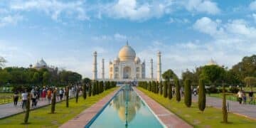 People walking along a pathway leading to the Taj Mahal, an iconic white marble mausoleum, surrounded by gardens and reflecting pools under a blue sky