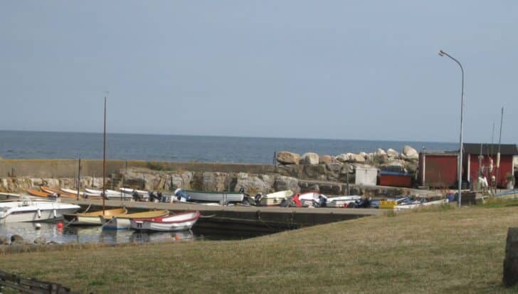 Small harbor in the town of Brantevik with several boats docked near a rocky shoreline, buildings to the right, and the ocean in the background.