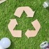 Recycling symbol made of cardboard surrounded by a paper cup, plastic bottle, glass bottle, light bulb, and aluminum can on grass