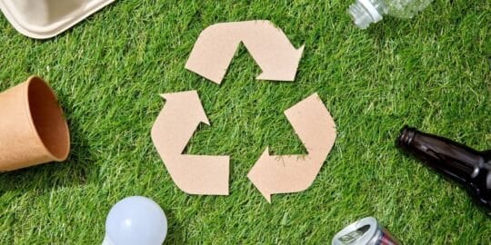Recycling symbol made of cardboard surrounded by a paper cup, plastic bottle, glass bottle, light bulb, and aluminum can on grass