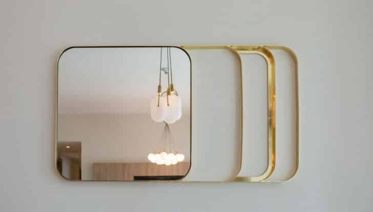 A modern mirror with a golden frame reflects a chandelier and part of a room, highlighting mirrors as non-recyclable items