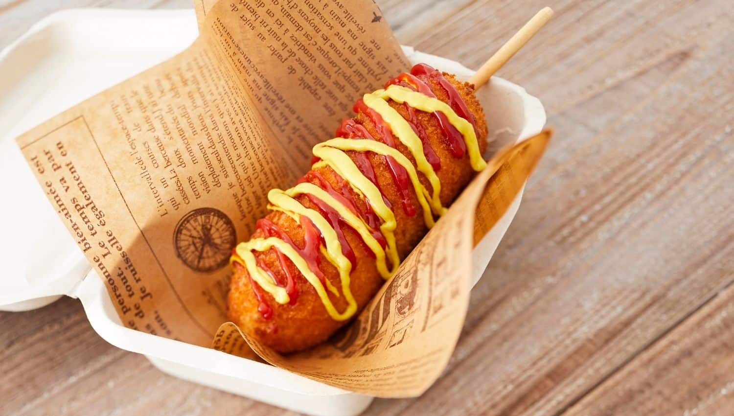 Corn dog with ketchup and mustard drizzle on a stick, wrapped in newspaper-style paper, in a takeout container on a wooden surface