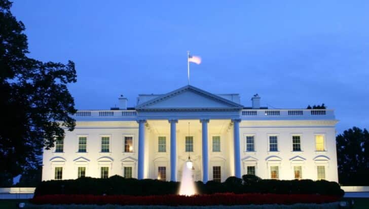 The White House illuminated against a twilight sky, iconic government building with American flag on top, surrounded by trees and gardens