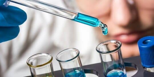 Close-up of a person in safety goggles using a pipette to add blue liquid to test tubes, likely related to significant chemical reactions