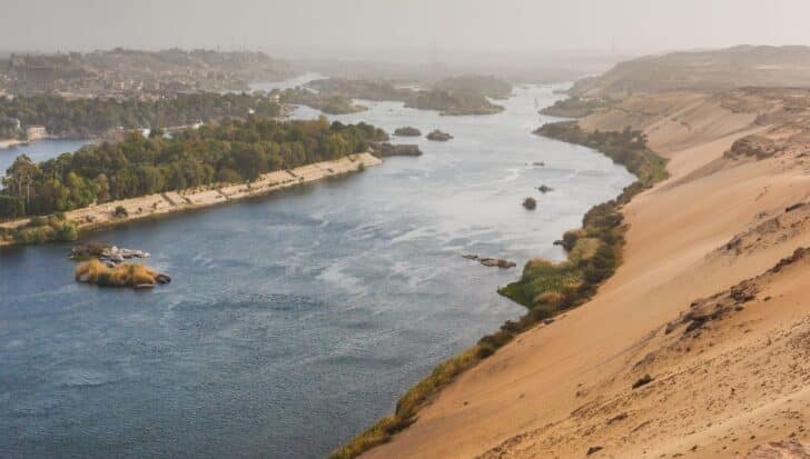 A wide view of the Nile River, with lush greenery along its banks and arid desert terrain on one side, showing the environmental context related to ancient Egyptian construction