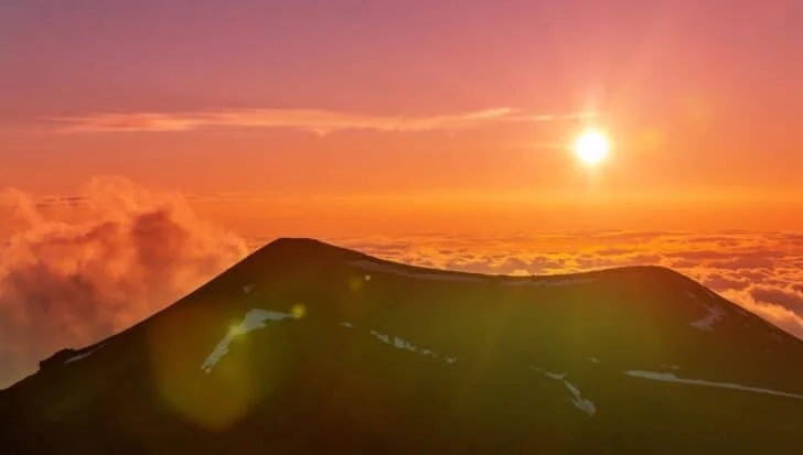 Silhouette of Mauna Kea against a vibrant sunset with clouds below its peak