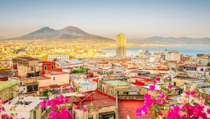 Urban landscape with colorful buildings, flowers, and a distant view of Mount Vesuvius in the background near the coast
