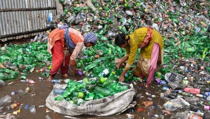 Two individuals sorting through a large pile of plastic bottles in a muddy area
