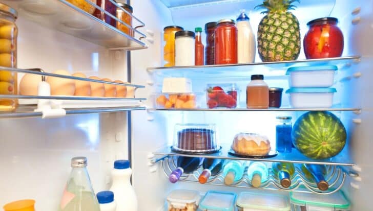 Open refrigerator filled with various foods including eggs, jars, a pineapple, watermelon, bottles, and containers.
