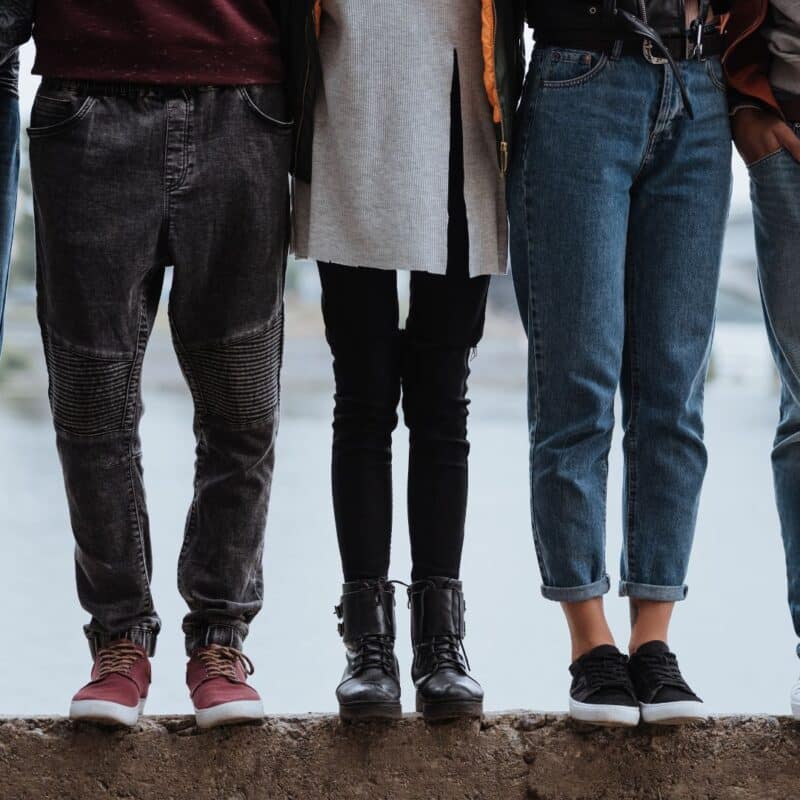 Four people standing outdoors with their legs visible, wearing casual clothing including jeans and boots