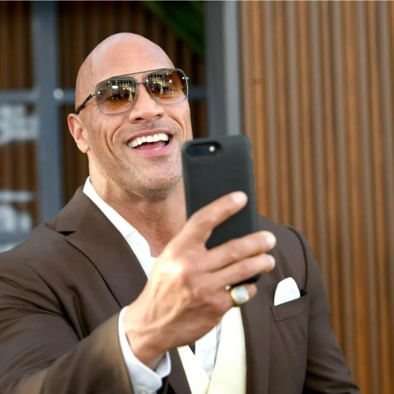 Dwayne Johnson taking a selfie with a smartphone, wearing sunglasses and a brown suit