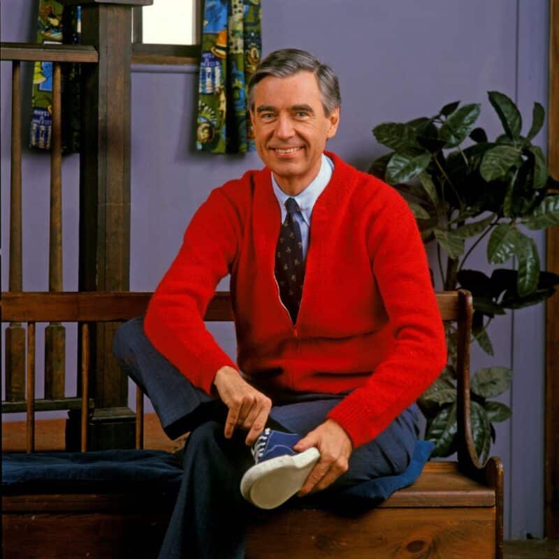 Mister Rogers is sitting on a wooden bench, wearing a hand-knitted red sweater