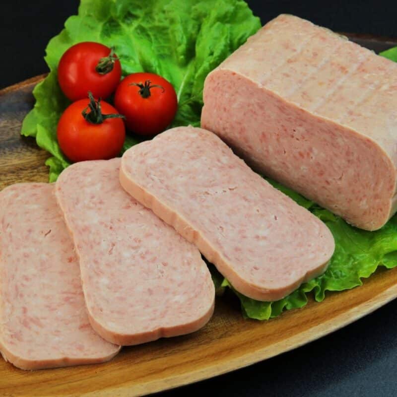 Slices and a block of Spam on a bed of lettuce leaves, accompanied by three whole tomatoes, on a wooden platter.