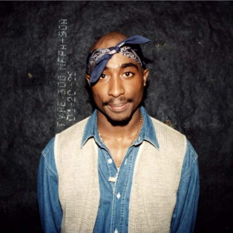 Tupac wearing a denim shirt and beige vest with a blue bandana on his head, standing against a dark textured background