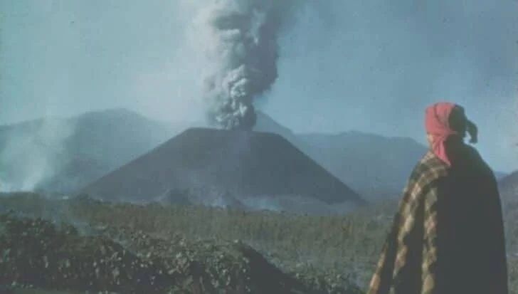 Person in a plaid garment observing an erupting volcano emitting smoke against a mountainous backdrop