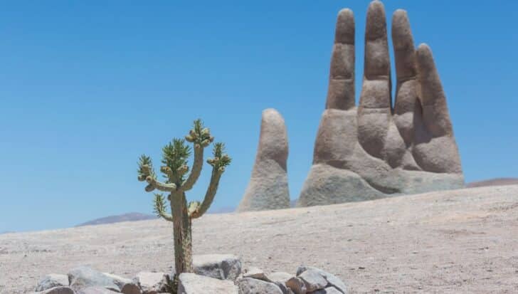 A large hand sculpture emerging from the desert sands with a small cactus in the foreground