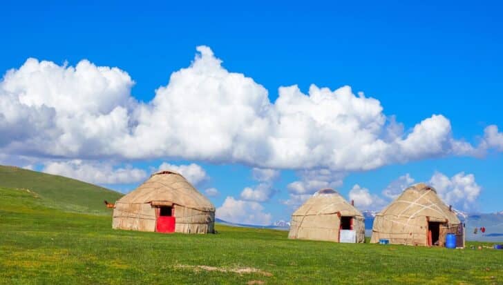 Three traditional yurts under a blue sky with clouds, located on a grassy plain in Kyrgyzstan