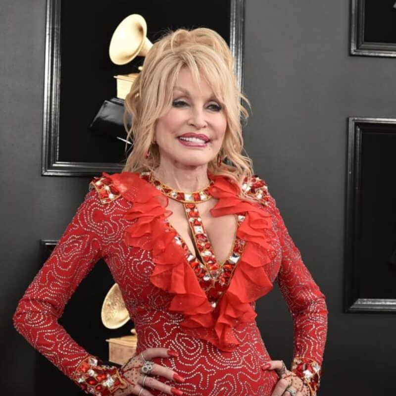 Dolly Parton in a red, ruffle-trimmed dress, standing in front of Grammy Award trophies