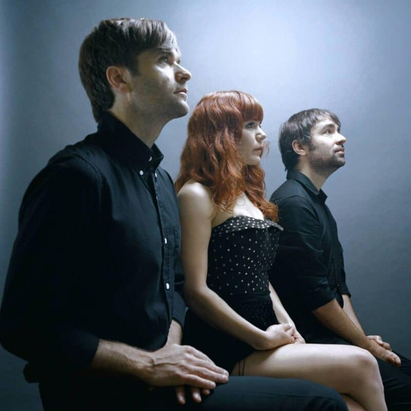 Three members of The Postal Service sitting against a minimalist backdrop, dressed in dark clothing