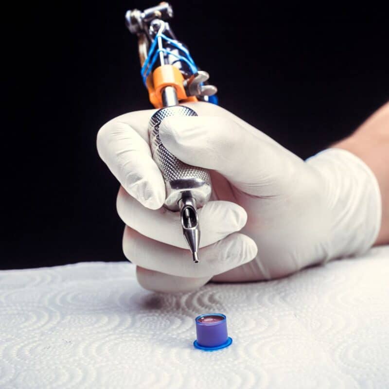 Hand in a white glove holding a modern tattoo machine over a small ink cap