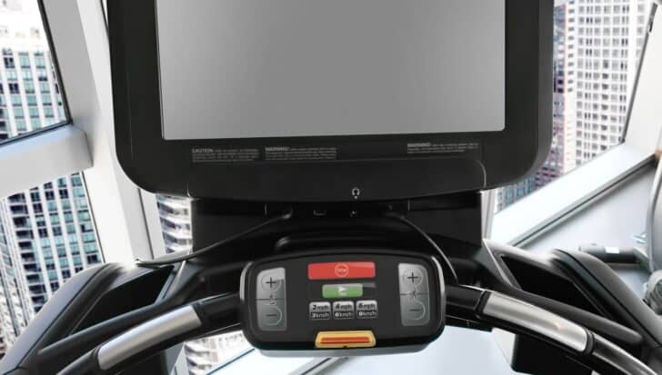 Treadmill display console with speed controls and stop button, positioned by large windows with city building views