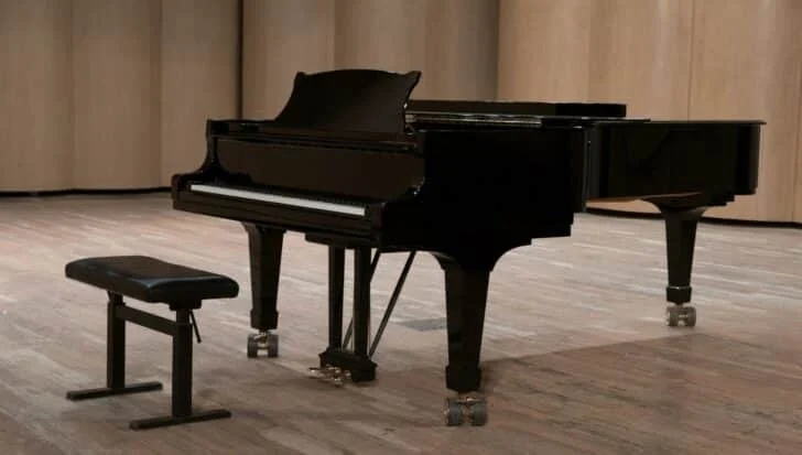 A grand piano on a wooden stage with a black piano bench, emphasizing the size and weight similar to cars
