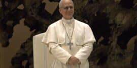 Pope Leo XIV, standing with hands folded, against a dark textured background