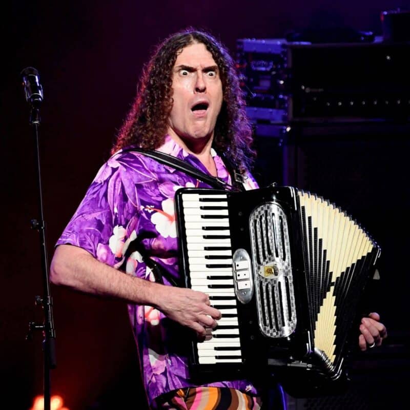 Weird Al Yankovic playing the accordion on stage, wearing a vibrant purple floral shirt