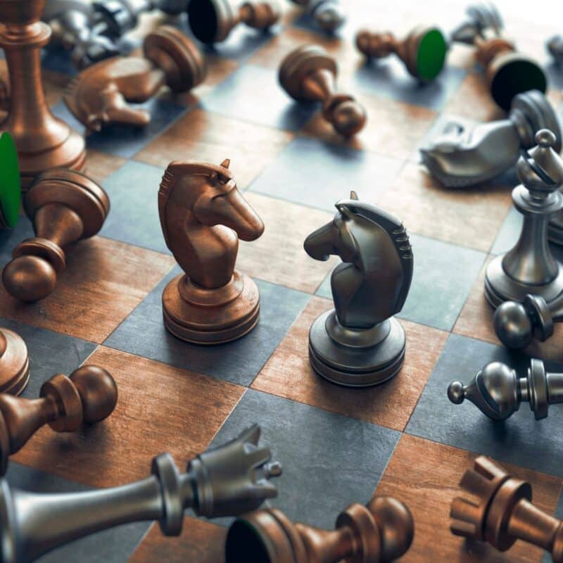 Wooden and metallic chess knights face each other on a chessboard surrounded by fallen pieces