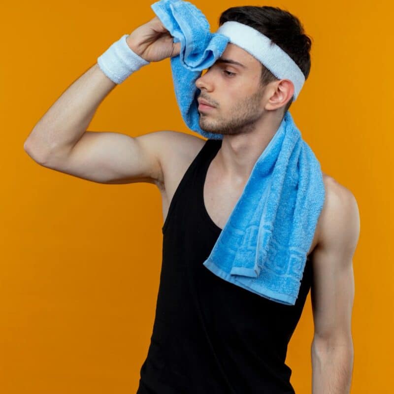 Young man wearing a black tank top, white headband, and wristband wipes sweat from his forehead with a blue towel