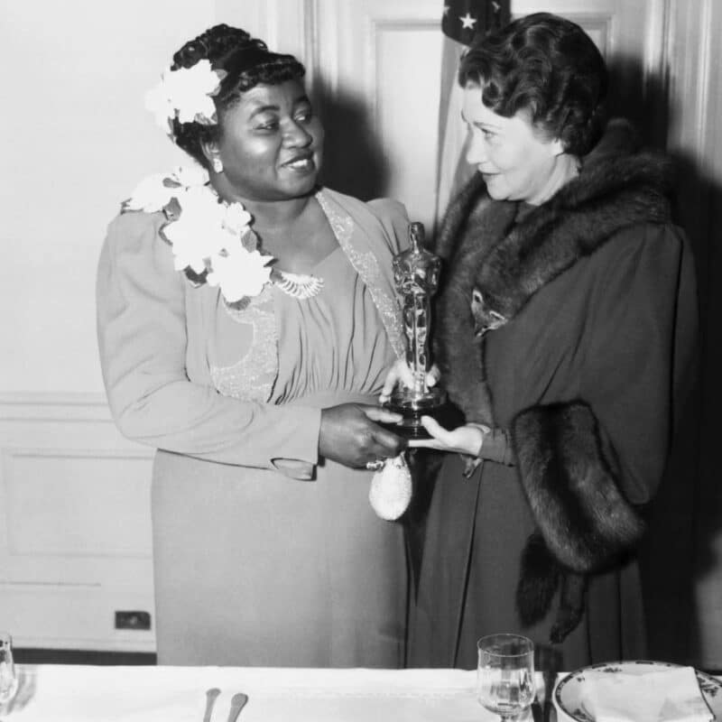 Hattie McDaniel, wearing a floral dress, smiles and holds her Oscar statuette